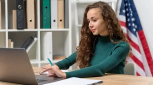 aluna estudando inglês avançado no notebook com bandeira americana e livros no fundo